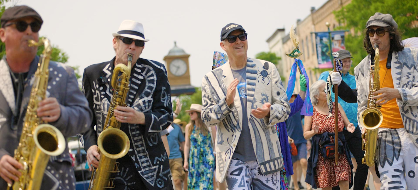 Three men in suits play saxophones and another man claps while they are marching in a parade.